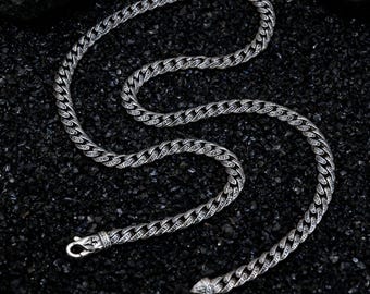 May include: A silver-colored curb chain necklace with a textured, interwoven design. The necklace is displayed on a dark, textured surface, showcasing its intricate details and the secure clasp.
