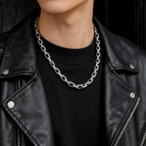 May include: A silver chain necklace with rectangular links. The necklace is worn over a black crew neck top and is paired with a black leather jacket. The image is a close-up shot of the necklace and upper body.
