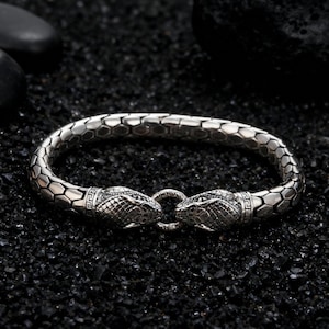 May include: Silver bracelet with a snake design. The bracelet has a textured, hexagonal pattern and two snake heads facing each other, connected by a ring clasp. The snakes have detailed scales and decorative patterns.