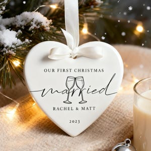 May include: A white heart-shaped Christmas ornament with the text "OUR FIRST CHRISTMAS" and "married" in cursive. Below the word "married" are two champagne glasses. The names "RACHEL & MATT" and the year "2023" are also printed on the ornament.