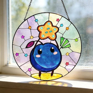 May include: A circular stained glass suncatcher featuring a blue character with a smiling face, holding a yellow star above its head. The background consists of pastel purple, yellow, and green glass segments decorated with small colorful flower accents.