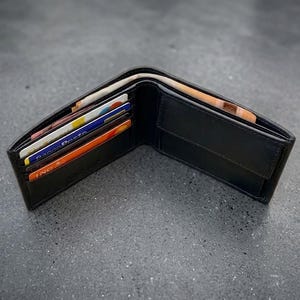May include: Open black leather wallet containing credit cards and banknotes. The wallet is shown open, revealing the internal sections and contents. The wallet is on a dark grey surface.