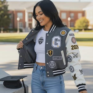 May include: A gray and white letterman jacket with multiple patches, including a large "G" and the year "2024". The jacket has black and white striped cuffs and collar. The person is wearing a white t-shirt and blue jeans.