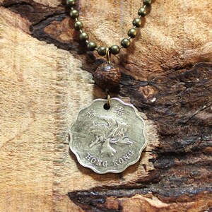 May include: A silver coin pendant featuring the words "HONG KONG" and Chinese characters, suspended from a brass-coloured beaded chain. A textured bead sits between the coin and the chain.