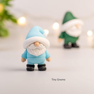 May include: A collection of miniature gnome figurines. The central gnome is light blue with a white beard and hat. Two other gnomes are in the background, one green and one blue. The text "Tiny Gnome" is at the bottom.