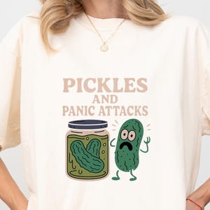 May include: A cream-coloured t-shirt with the words "Pickles and Panic Attacks" in brown. The design shows a jar of pickles and a cartoon pickle with a worried face.