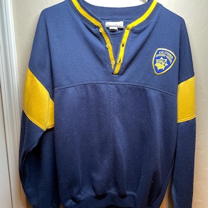Op de afbeelding: Marineblauw sweatshirt met gele accenten op de kraag, mouwen en schouders. Het heeft een knoopsluiting en een patch met de tekst "CALIFORNIA HIGHWAY PATROL". Het sweatshirt hangt aan een zwarte hanger.