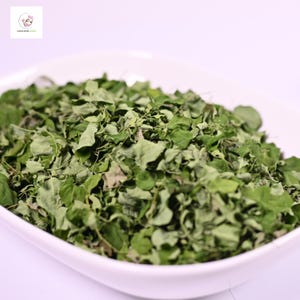 May include: A white, oval bowl overflowing with dried moringa leaves. The leaves are various shades of green, with some showing signs of drying. The image is well-lit, highlighting the texture and colour of the leaves.