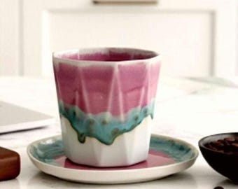 May include: A pink and teal ceramic coffee cup with a matching saucer. The cup has a geometric design and a white base. The saucer is pink with a teal rim. A small black bowl of coffee beans sits nearby.