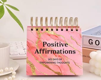 May include: A pink desk calendar with gold accents and a marble design. The calendar reads "Positive Affirmations" and "365 Days of Empowering Thoughts". The calendar is spiral-bound with gold metal rings.