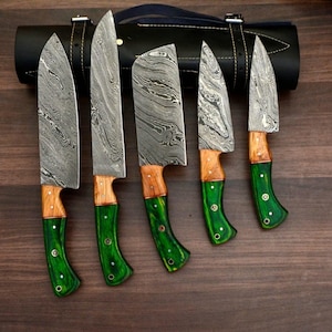 Può includere: Set di cinque coltelli da cucina in acciaio Damasco con manici in legno e verdi. Le lame presentano un motivo unico. È visibile un rotolo di coltelli in pelle nera.