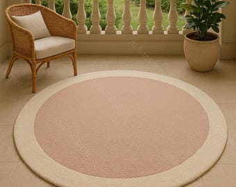 May include: A round rug with a light pink center and a cream-colored border. The rug is placed on a tiled surface, with a wicker chair and a potted plant visible in the background. The rug is designed for indoor or outdoor use.