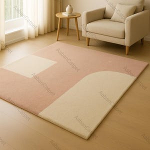 May include: A rectangular rug with a geometric pattern in a living room setting. The rug has a pink base with beige and off-white shapes. It's placed on a wooden floor, with a beige armchair and side table visible in the background.
