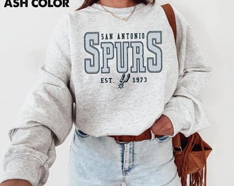 May include: Ash-colored sweatshirt featuring the text "SAN ANTONIO SPURS" in a light blue, outlined font. Below the text is "EST. 1973" with a spur logo. The long-sleeved top is made of a soft material.