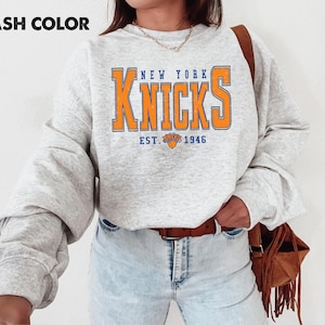 May include: Ash color sweatshirt with the text "NEW YORK KNICKS" in orange and blue. The sweatshirt has a crew neck and long sleeves. The text "EST. 1946" is below the team name, with a basketball graphic.