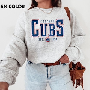 May include: Ash color sweatshirt with the text "CHICAGO CUBS EST. 1870" in blue and red lettering. The sweatshirt is a light gray color and has long sleeves. A brown leather purse with fringe is on the right side.