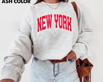 May include: Ash color sweatshirt with the text "NEW YORK" in red block letters. The long-sleeved top is paired with light blue jeans and a brown leather belt. A brown leather purse with fringe is visible.