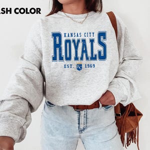 May include: Ash-colored sweatshirt featuring the text "Kansas City Royals" in blue, with "EST. 1969" below. The sweatshirt has long sleeves and a crew neck. The image shows a close-up of the upper body.