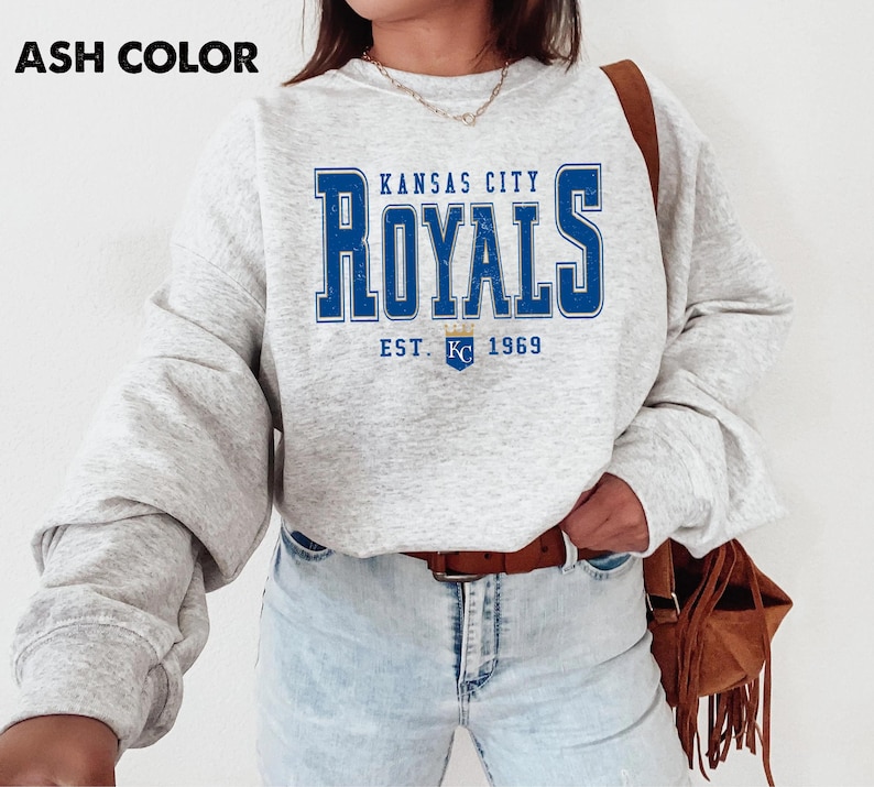 Pu&ograve; includere: Felpa grigio chiaro con la scritta blu "Kansas City Royals" e "EST. 1969" sotto. La felpa ha maniche lunghe e girocollo. L'immagine mostra un primo piano della parte superiore del corpo.
