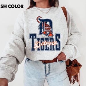 May include: Ash color sweatshirt featuring the Detroit Tigers logo. The logo includes a tiger graphic inside a navy blue "D" and the word "Tigers" in navy blue with an orange script font. The sweatshirt has long sleeves.