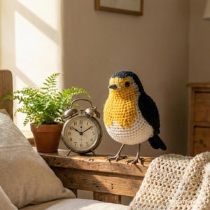 May include: A handcrafted, crochet bird with a yellow breast, black wings, and a white belly. The bird is perched on a wooden surface next to a vintage alarm clock and a potted fern. The clock shows the time as 10:09.