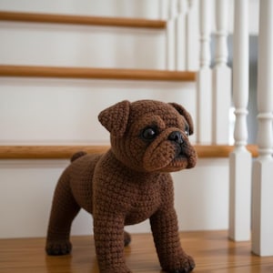 Pode incluir: Uma estatueta de buldogue de crochê marrom. O cão feito à mão tem um focinho curto, orelhas caídas e uma cauda pequena. O cão está em pé sobre uma superfície de madeira em frente a uma escada com balaústres brancos e um corrimão de madeira.