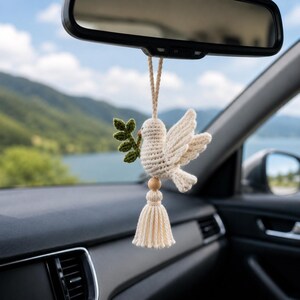 May include: A cream-colored, crocheted dove car charm with a green olive branch in its beak. The dove hangs from a string, with a wooden bead and tassel below. The charm is suspended from a car's rearview mirror.