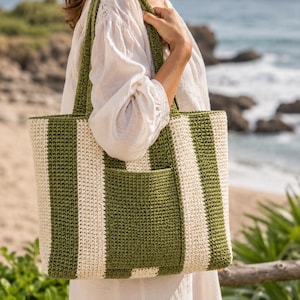 Könnte beinhalten: Eine gehäkelte Tragetasche mit grün-weißen Streifen, einer Vordertasche und passenden Trägern. Die Tasche wird von einer Person getragen, die ein weißes Kleid trägt, mit einem Strand und Meer im Hintergrund.