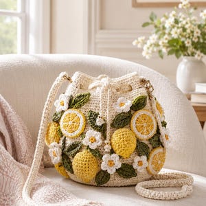 May include: Cream crochet handbag decorated with yellow lemons, white flowers, and green leaves. The bag features a drawstring closure, a long strap, and a smaller handle. The bag is on a white chair.