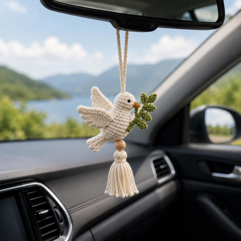 May include: A crocheted dove car charm, cream-colored with a green olive branch in its beak, hangs from a car's rearview mirror. A wooden bead and tassel complete the design. The background shows a lake and mountains.
