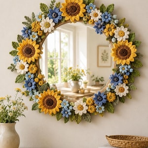 May include: A round wall mirror framed with handcrafted crochet flowers and leaves. The floral arrangement features yellow sunflowers, white daisies, and small blue blossoms interspersed with green foliage, creating a textured border.