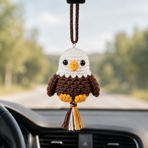 Può includere: Un ciondolo per auto a forma di aquila calva all'uncinetto. L'aquila ha la testa bianca, il corpo marrone e il becco e le zampe gialle. È appeso a un cordoncino marrone e ha un nappa di filato marrone, bianco e giallo.