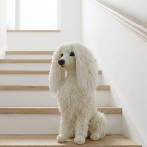 Może przedstawiać: Biała, puszysta rzeźba psa pudla siedzi na drewnianych schodach. Pies ma teksturowane, szorstkie futro i ciemne oczy. Tło przedstawia białą ścianę i okno na szczycie schodów.