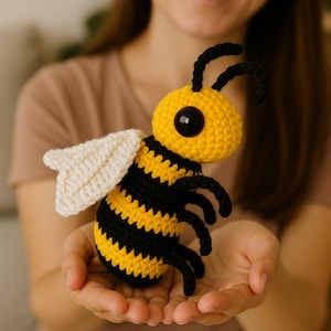 May include: A handmade crochet bee toy. The bee features a yellow body with black stripes, large black eyes, and black antennae. It has white wings and black legs. The toy is held in open hands.