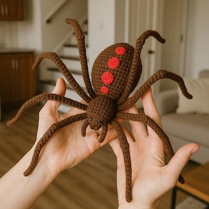 May include: A brown crochet spider with eight legs and red circular markings on its back. The spider is held in open hands, highlighting its detailed craftsmanship. This plush toy is a unique decorative item.