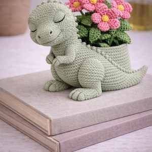 May include: A crocheted dinosaur planter in soft green holds pink and yellow crocheted flowers. The dinosaur is sitting on top of two books. The planter is a charming, handmade item, perfect for adding a touch of whimsy to any space.