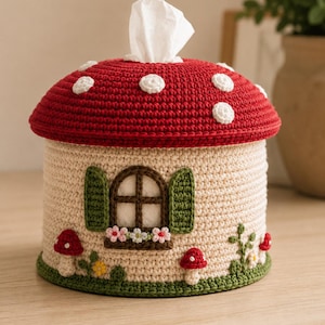May include: A cylindrical crochet tissue box cover shaped like a mushroom house. The top is a red cap with white spots, while the base is beige with a brown window, green shutters, small flowers, and red mushroom accents on a green border.