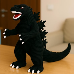 Puede incluir: Una figura de Godzilla negra tejida a ganchillo con garras y dientes blancos y ojos rojos. El juguete hecho a mano está de pie con la boca abierta, mostrando un interior rojo. La figura tiene una larga cola y aletas dorsales puntiagudas.