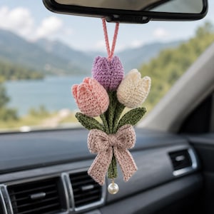 May include: A crocheted car hanging ornament featuring three tulips in pastel pink, purple, and cream colors. The flowers have green stems and leaves, tied together with a light pink crocheted bow and finished with a single white pearl bead at the base.