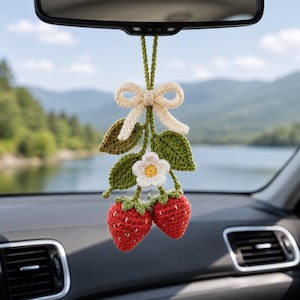 May include: Crocheted strawberry car charm with red berries, green leaves, and a white flower. The charm is suspended from a beige bow and string, designed to hang from a car's rearview mirror. A charming, handmade accessory.