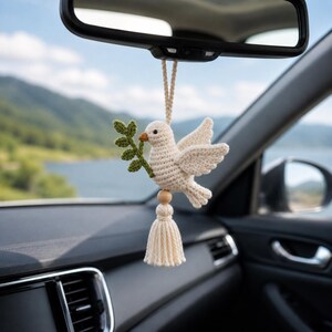 May include: A cream-colored, crocheted dove car charm with a green olive branch. The dove is suspended from a rearview mirror, with a wooden bead and tassel below. The background shows a scenic view of a lake and mountains.