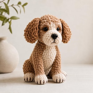 May include: A crocheted dog figurine in shades of brown and cream. The handmade puppy has floppy ears, a white chest, and black eyes and nose. The dog is sitting, with its paws in front. The background is a neutral color.