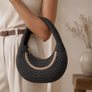 May include: A crescent-shaped handbag crafted from dark charcoal woven material, featuring a single integrated handle and a prominent curved accent stripe in a lighter tan color along the front. The bag includes a gold-toned zipper closure.