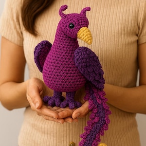 May include: A handmade crochet bird in shades of purple and yellow. The bird has a detailed tail, wings, and feet. The bird is held in open hands. The bird's beak is yellow, and the body is a deep purple.