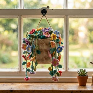 Könnte beinhalten: Ein hängender Häkelkorb gefüllt mit bunten Häkelblumen in Rot-, Gelb-, Blau- und Lilatönen. Der Korb hängt vor einem Fenster, mit einer kleinen Sukkulente und einem Messingzerstäuber auf der Fensterbank.