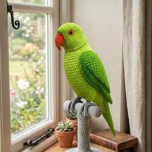 May include: A handcrafted crochet parrot, predominantly green with an orange beak and yellow eyes, perched on a grey crochet stand. The bird is positioned on a wooden surface near a window, overlooking a garden.