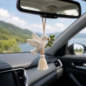 May include: A crocheted dove car charm, holding a green olive branch in its beak. The dove is cream-colored, with a wooden bead and tassel below. The charm hangs from a string, suspended in front of a car's rearview mirror, with a scenic background.