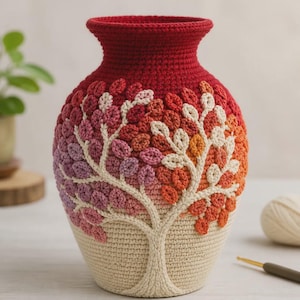May include: A handcrafted crochet vase in a warm color palette. The vase features a tree design with leaves in shades of red, orange, and pink, transitioning to a beige base. The top is a deep red, and the vase is likely made of yarn.