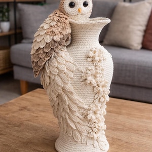 May include: A handcrafted, cream-colored crochet vase with a detailed owl perched on the side. The owl has brown and beige feathers, and the vase is adorned with floral embellishments. The vase is displayed on a wooden table.