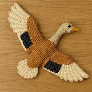 Może przedstawiać: Ręcznie robiona szydełkowa kaczka w locie, z beżową głową i skrzydłami, brązowym korpusem i czarnymi akcentami na skrzydłach. Kaczka jest prezentowana na drewnianym tle, ukazując jej szczegółowe wykonanie.